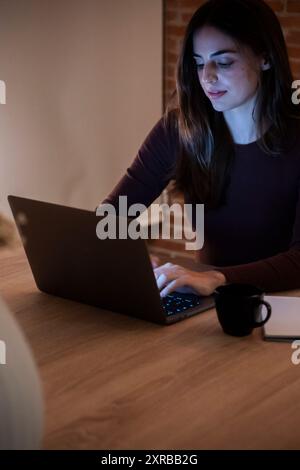 Concentrated young woman working late in home office, using laptop, copy space. Casual girl in the light of the screen on the background of the illuminated room. Technology and work concept Stock Photo