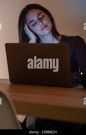 Tired businesswoman working on computer until night. Portrait of a stressed casual woman with a headache at the desk. Exhausted businesswoman working late at night on laptop in office or home. Stock Photo