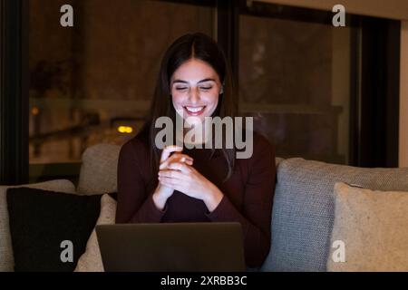 Woman sitting on sofa at night relaxes watching funny videos on her laptop. Pretty female with long hair smiles while she watches funny movie using computer connection. Dating online people. Overtime on notebook on sofa Stock Photo