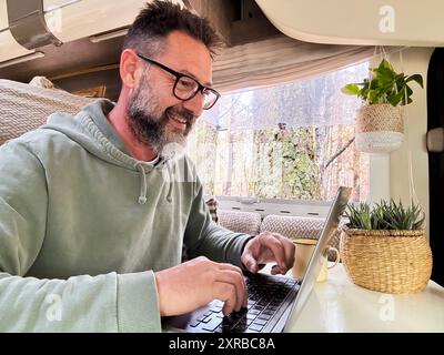 Man working inside a camper van in modern online job business. Remote worker content creator influencer vanlife lifestyle concept people. Modern mature male using computer in indoor leisure activity Stock Photo