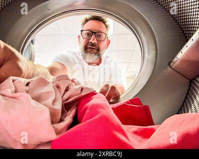 Man Doing Laundry Reaching Inside Washing Machine. One adult male washing clothes. View from inside the laundry. Homework husband. People using washing machine in laudromat Stock Photo