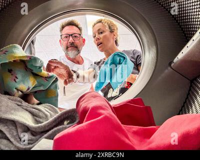 Young couple doing laundry View from the inside of washing machine.Happy adult man and woman cleaning and washing clothes together in homework or business activity. Using laudromat Stock Photo