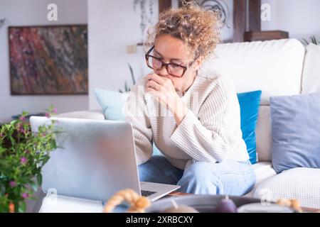 Thoughtful woman holding laptop on laps, pondering ideas or tasks, sitting on couch at home, dreamy young female touching face and looking the display, lost in thoughts, waiting for message online Stock Photo