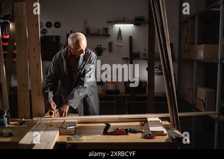 70s carpenter using a tape measure to measure wood for cut. Elderly man make furniture in the carpentry workshop Stock Photo