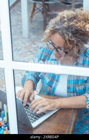 Woman in checked shirt working on laptop while sitting at desk near window in a laboratory office at home. Vision through the window. Small new business female people lifestyle using computer online Stock Photo
