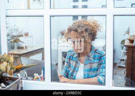 Woman in checked shirt working on laptop while sitting at desk near window in a laboratory office at home. Vision through the window. Small new business female people lifestyle using computer online Stock Photo