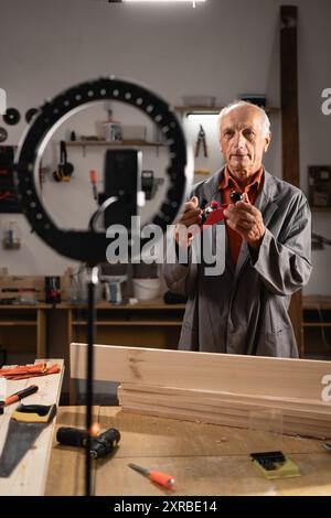 Elderly carpenter with phone on tripod recording vlog, communicating with followers, livestreaming for carpentry blog in social media working in his Stock Photo