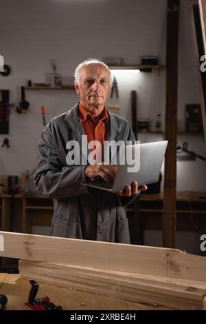 Portrait of an elderly male carpenter standing in his workshop using a laptop computer. Woodworking and small business Stock Photo