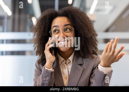 Young African American businesswoman with glasses smiling while talking on phone. Confident professional at workplace inside office. Positive and successful representation of women in business. Stock Photo