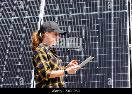 Female maintenance worker wearing plaid shirt and cap analyzing the performance of solar panels using a digital tablet. Stock Photo