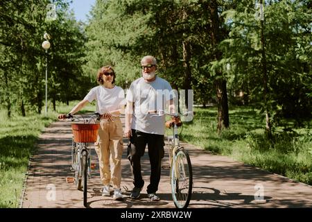 Senior loving couple dressed in casual clothes and wearing sunglasses having bike walk in park during warm summer day Stock Photo