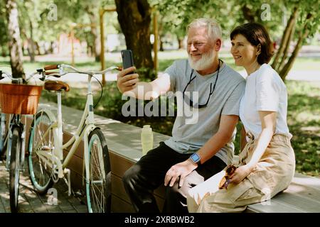 Senior couple dressed in casual outfit sitting on bench while taking selfie Stock Photo