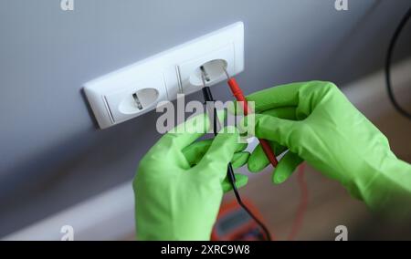 Hands in green gloves plug voltmeter into an outlet in wall Stock Photo