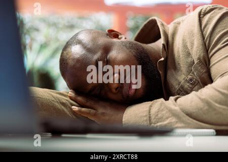 Successful businessman sleeping on his home desk while he works on laptop, taking a nap after suffering from fatigue and tension. Exhausted freelancer falls asleep from burnout. Stock Photo