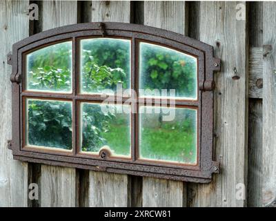 Rusty wrought iron window frame with mirror glass on a shed wall, reflecting garden and sky. A touch of nostalgia and past beauty. Stock Photo