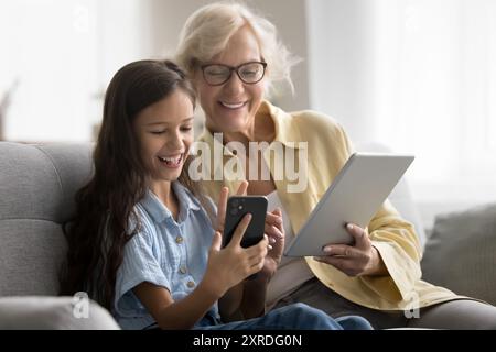 Cheerful grandma and granddaughter kid using digital gadgets Stock Photo