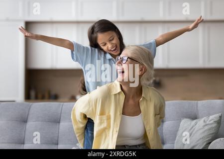 Happy excited strong grandma piggybacking granddaughter, sitting on home sofa Stock Photo