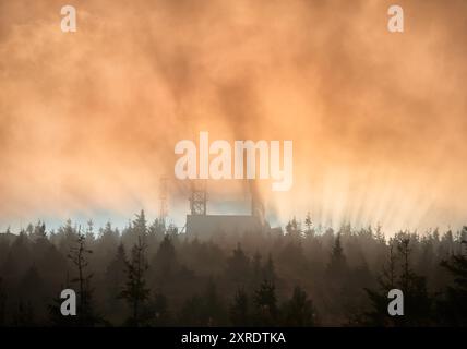 Sun rises, its rays pierce through the mist, casting dramatic beams of light over landscape. Antenna tower and building emerge from fog, surrounded by dense forest of pine trees. Stock Photo