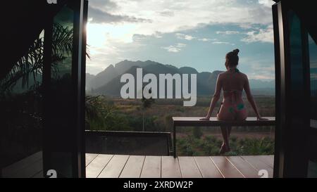 Woman in bikini sits wooden balcony looking sunset mountain range in Thailand resort. Girl relax in holiday exotic wild nature hut villa. Outdoor lifestyle travel on summer holiday vacation back view Stock Photo