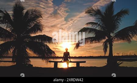 Watching the sunset by the sea, a woman sits on a bench between palm trees, surrounded by nature's beauty in a tranquil tropical paradise Stock Photo