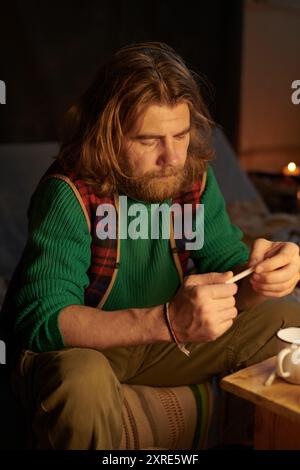 Man with long hair and beard concentrating while inspecting small object in his hands, sitting on patterned couch in cozy indoor setting with dim lighting Stock Photo