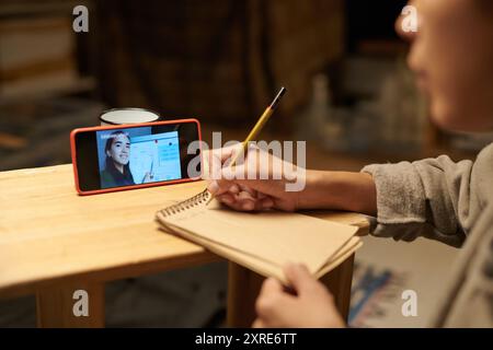 Person taking notes while attending an online lecture on a bright tablet screen on a wooden table, holding a pencil, focused on learning Stock Photo
