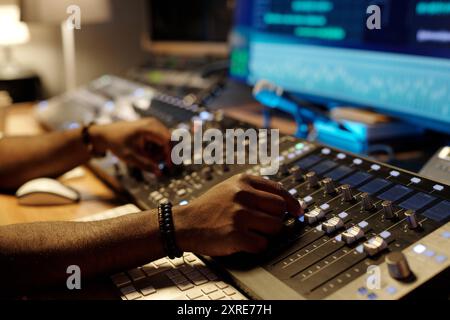 Close-up view of person adjusting audio levels on professional mixing console in recording studio setting with multiple screens displaying audio waveforms and tracks Stock Photo