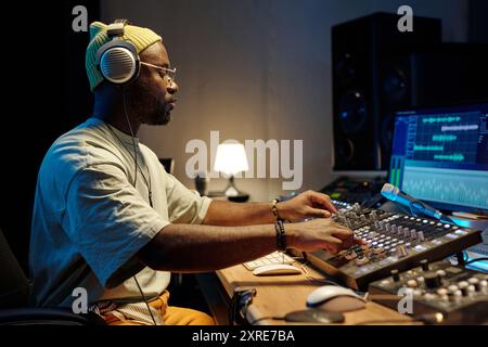 African American man wearing headphones working on music production in modern studio setting. Filled with electronic devices and monitors showcasing editing software Stock Photo