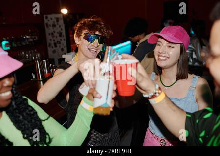 Group of young cheerful intercultural friends clinking with cups of soda and bottles of beer while toasting at party in club Stock Photo