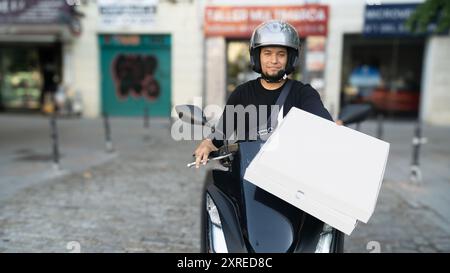 A man wearing a helmet is holding a pizza box on a motorcycle. Concept of adventure and excitement, as the man is ready to deliver the pizza to his cu Stock Photo
