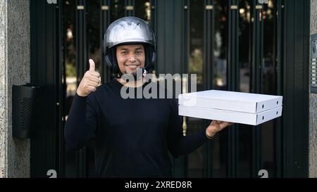 A man wearing a helmet is holding a pizza box and giving a thumbs up. Concept of excitement and satisfaction, as the man is happy to be delivering the Stock Photo