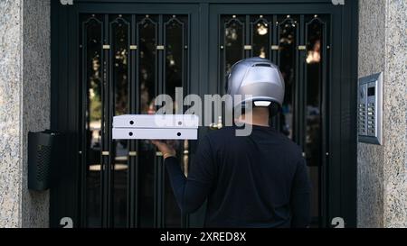 A man wearing a helmet is holding two boxes of pizza. The scene is set outside a building with a metal door Stock Photo