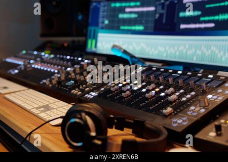 Professional audio mixing board with multiple channels illuminated, showing various sound frequencies on screen in background with headphones resting on desk nearby Stock Photo