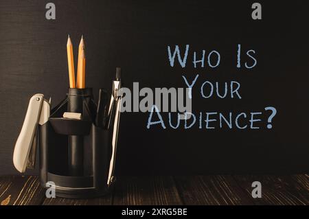 A pencil holder with two pencils and a marker sits on a wooden table. The words Who is your audience are written on the table Stock Photo