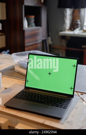 Displaying laptop with green screen placed on wooden desk in modern workspace featuring shelves with office supplies and window in background Stock Photo