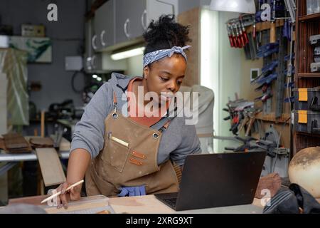 Craftswoman with a headband seen working in workshop using laptop while shaping wooden piece on workbench. Tools and equipment seen neatly organized in background Stock Photo