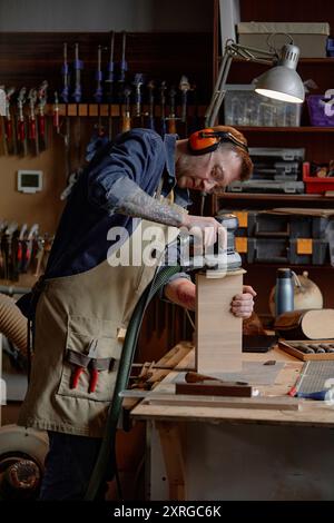 Man using electric sander on wooden block in workshop filled with tools and gadgets. Concentrating while standing at workbench in woodworking setting with shelves in background Stock Photo