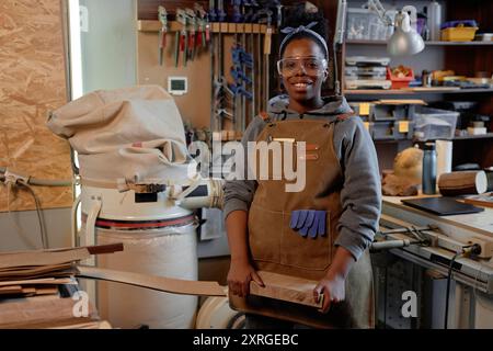 African American woman in workshop wearing safety gear and apron, sanding wooden piece with visible tools and equipment in background Stock Photo