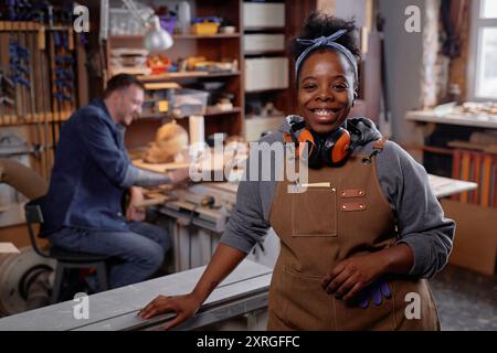 Portrait of happy female woodworker beaming in workshop wearing overalls and protective gear, with colleague working in background Stock Photo