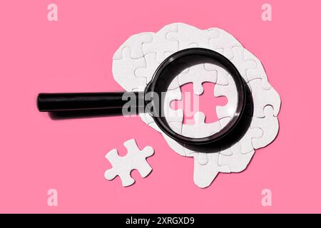 Artistic depiction of a brain composed of white puzzle pieces, highlighted by a magnifying glass. Final element is set aside on a pink background. Stock Photo