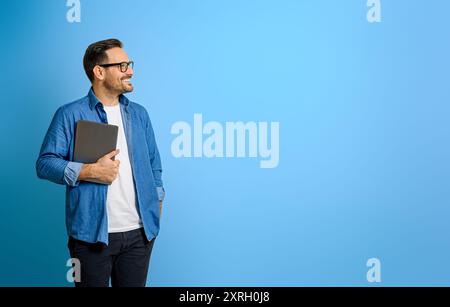 Thoughtful smiling businessman holding digital tablet and looking away against blue background Stock Photo