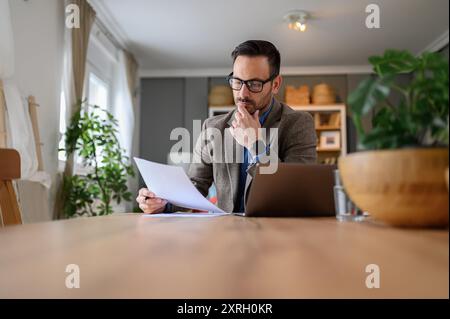 Focused businessman in suit reading documents with laptop on desk while working from home office Stock Photo