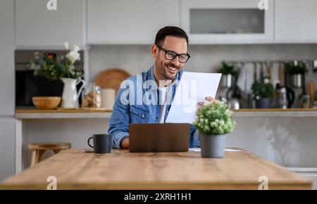 Smiling young businessman analyzing report and working over laptop on desk in home office Stock Photo