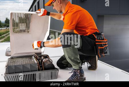 A technician in an orange shirt inspects and repairs a camper van rooftop HVAC unit while wearing gloves and safety gear. Stock Photo