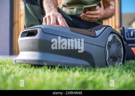 A person operates a robotic lawn mower with a smartphone while standing on lush green grass in their backyard. Stock Photo