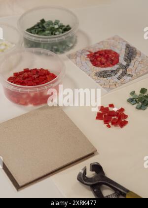 Tile nipper, mosaic base, out print of an antique pomegranate mosaic pattern, containers with red, green and white glass pieces on a white surface. Stock Photo
