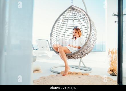 Beautiful middle-aged woman sitting in a comfortable hanging chair on open house terrace with a modern laptop working remotely at home office. Modern Stock Photo