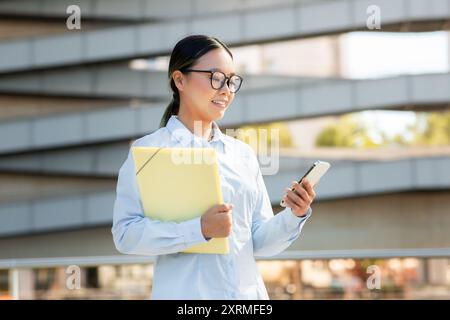 Young Professional Woman Smiling While Using Smartphone Near Modern Building Stock Photo