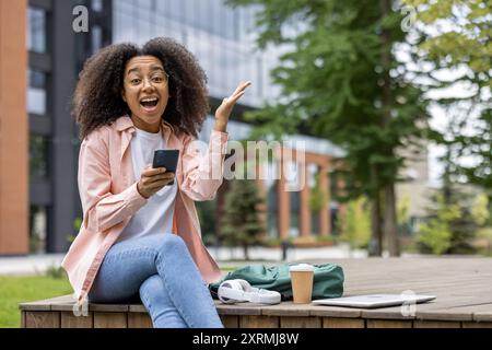 Cheerful African American student sitting outdoors on campus, using a smartphone and expressing excitement. The scene is full of vibrant energy and shows a relaxed, academic environment Stock Photo