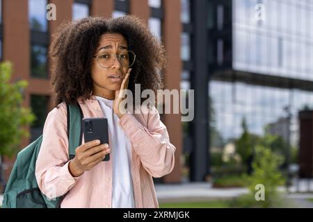 African American female student stands outdoors, looking concerned while holding a smartphone. The background features a modern campus building. The image conveys worry Stock Photo
